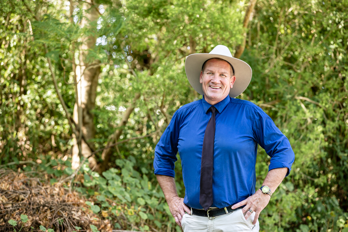 Adult standing outdoors in a leafy garden wearing a wide-brim hat, blue shirt, and tie