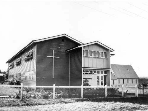 Black-and-white photo of an early school building with timber cladding, large cross on the wall, and a fence in the foreground