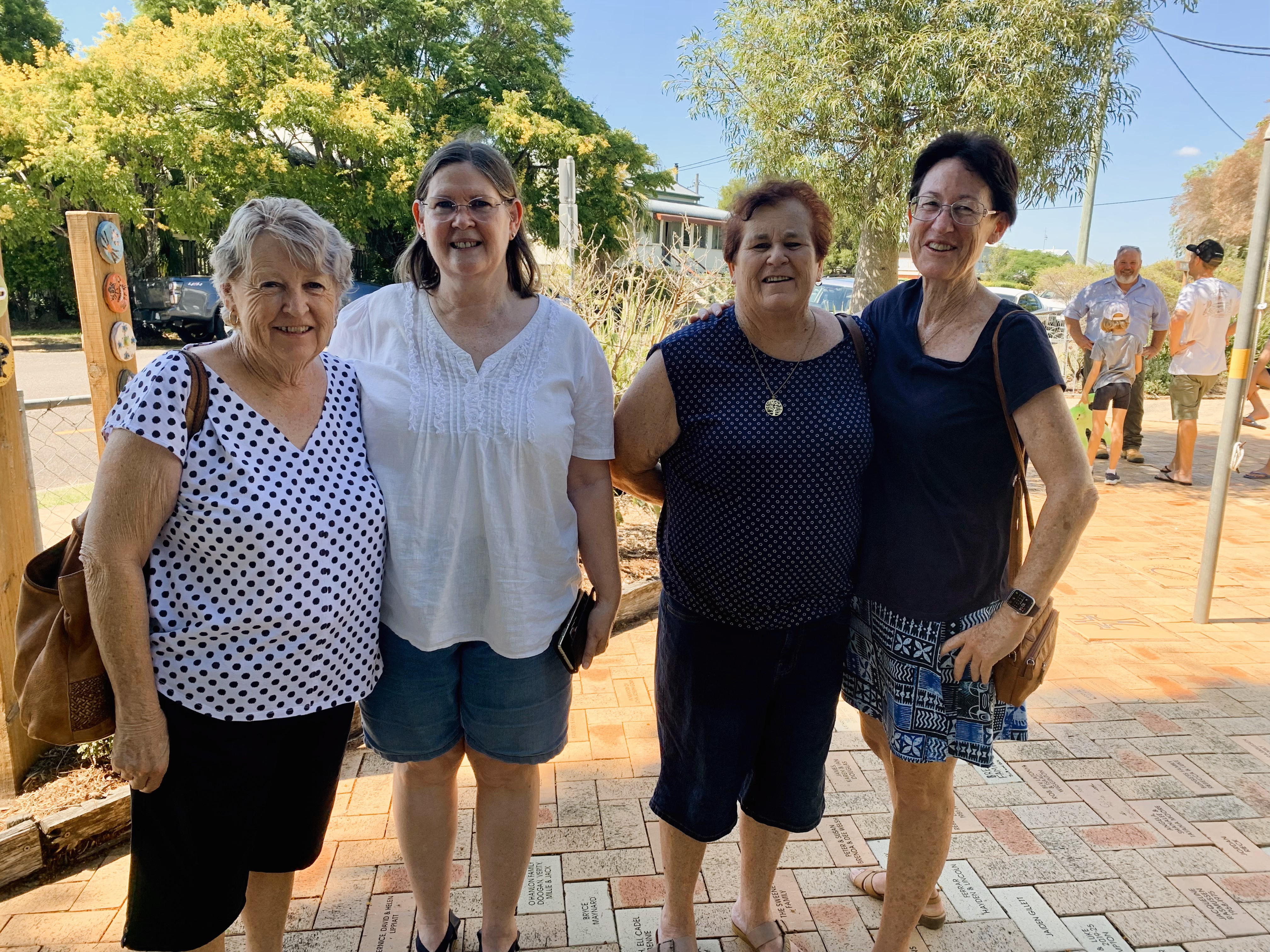 Four adults standing together outdoors on a paved courtyard at a school, with trees and buildings in the background