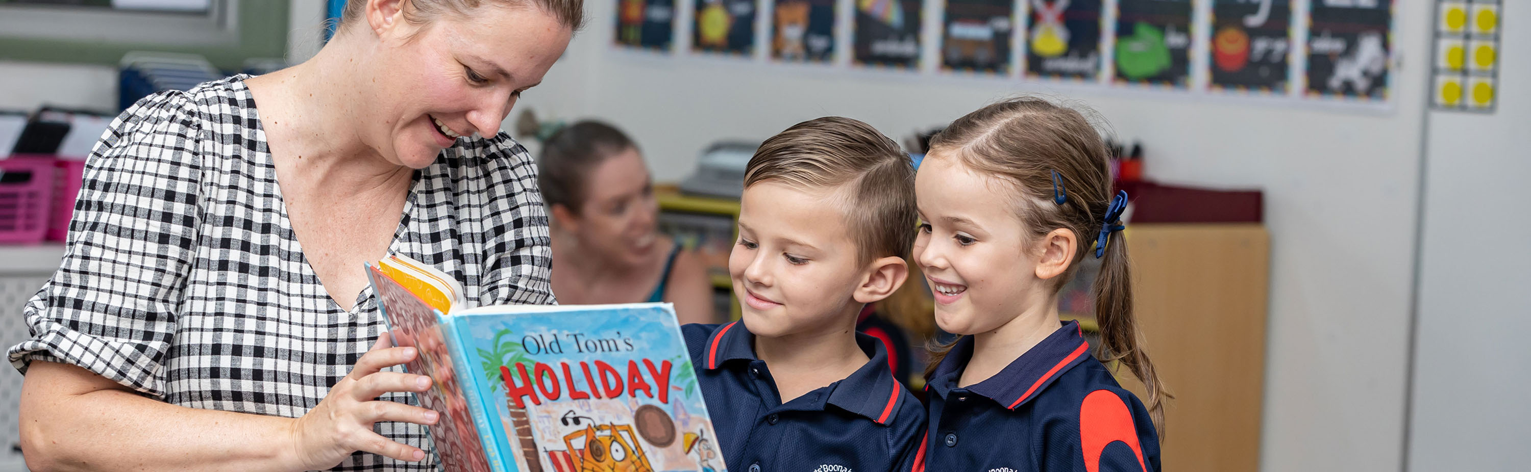 Teacher reading a picture book with two young students in a classroom, with colourful learning displays in the background
