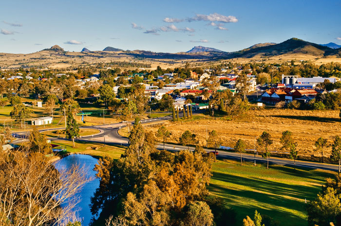 Rural town with houses, roads, and a river in the foreground, surrounded by dry hills and scattered trees under a blue sky